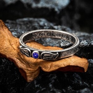 Op de afbeelding: Een zilveren ring met een verweerde afwerking, met een blauwe edelsteen en vleugelontwerpen. De ring heeft de inscriptie "Fly on my sweet angel". De ring wordt getoond op een bruin blad en een donkere stenen achtergrond.