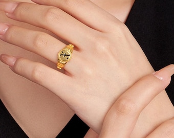 May include: A gold ring with an engraved design and a black letter "J" at the centre. The ring is on a finger, highlighting its intricate details and craftsmanship. The ring's band features a textured pattern.