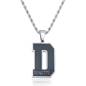 Könnte beinhalten: Eine silberfarbene Halskette mit einem großen, dunkelgrauen Anhänger in Form des Buchstaben "D". Der Anhänger hat einen silbernen Rand und der Name "DENETTE" ist unten aufgedruckt. Die Kette hat ein gedrehtes Seildesign.