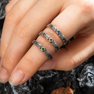 Op de afbeelding: Drie zilveren ringen met een vleugelontwerp en gekleurde edelstenen. De ringen hebben een donkere, geoxideerde afwerking en zijn versierd met groene, gele en blauwe edelstenen. De ringen worden op een vinger getoond.