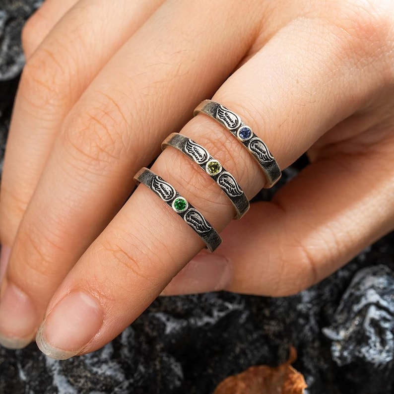 Op de afbeelding: Drie zilveren ringen met een vleugelontwerp en gekleurde edelstenen. De ringen hebben een donkere, geoxideerde afwerking en zijn versierd met groene, gele en blauwe edelstenen. De ringen worden op een vinger getoond.