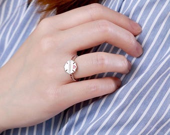 May include: A rose gold ring with a unique design. The ring features a small, white, shield-shaped centerpiece with a pink heart and text. The band is adorned with small, pink stones. The ring is on a person's finger, against a blue and white striped background.