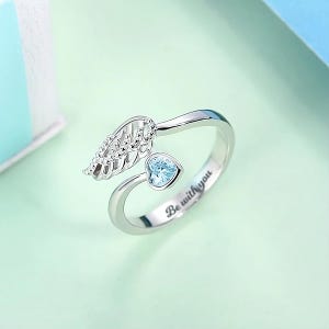 May include: Silver ring with a delicate wing design and a small blue heart-shaped stone. The band is inscribed with the words "Be with you."