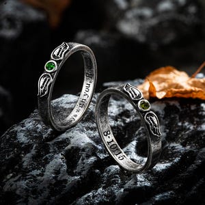Op de afbeelding: Twee zilveren ringen met een vintage afwerking, elk met een groene edelsteen en een vleugelontwerp. E&eacute;n ring heeft de inscriptie "always with you, Rod" en de andere "8-10-15".