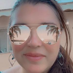 Puede incluir: Una mujer con gafas de sol de montura dorada con lentes espejados que reflejan palmeras y una escena de playa. Lleva una camisa verde y tiene el pelo largo y castaño.