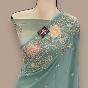 Op de afbeelding: Een turquoise stof met bloemenborduurwerk. Het borduurwerk heeft roze en perzik bloemen met groene bladeren. De stof is over een mannequin gedrapeerd. Een label met tekst is zichtbaar.