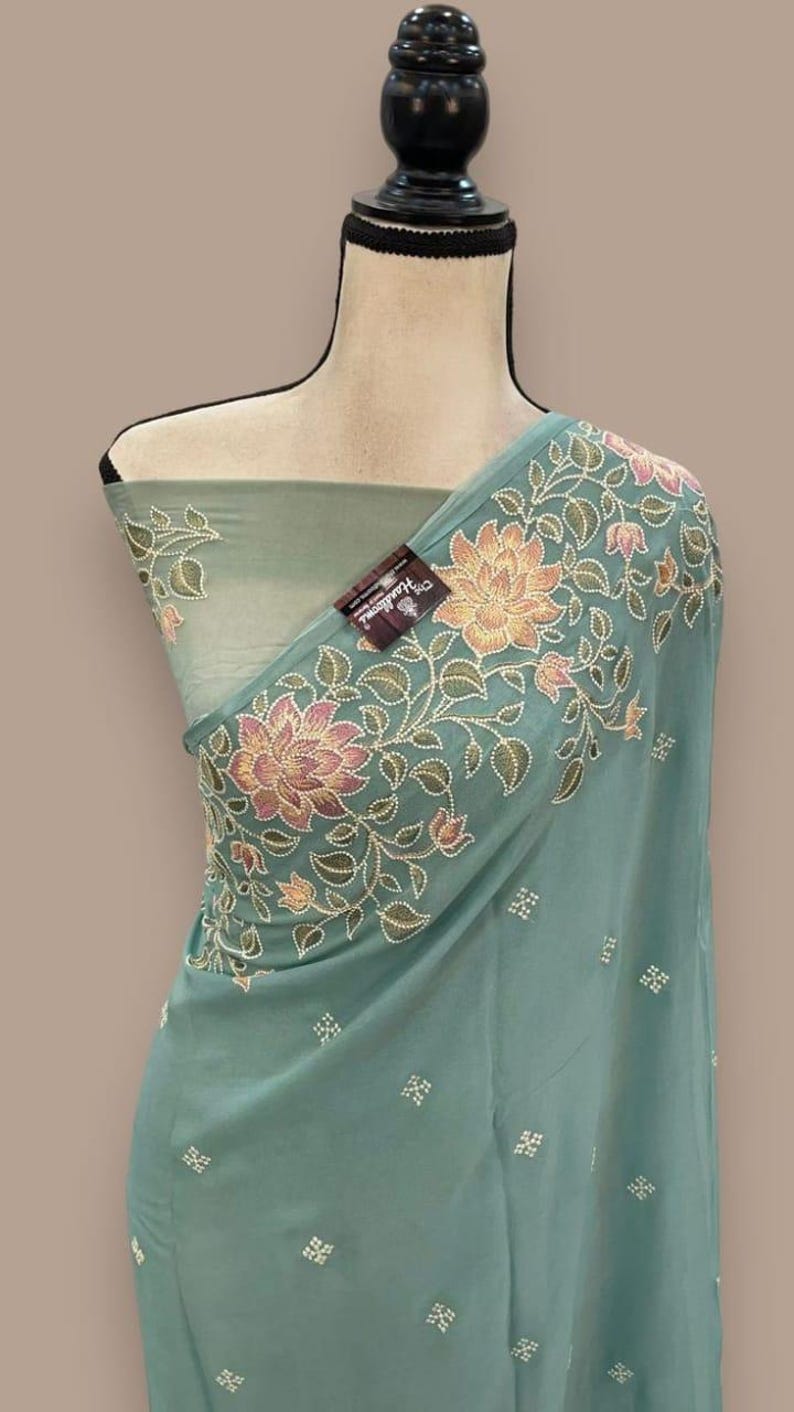 Op de afbeelding: Een turquoise stof met bloemenborduurwerk. Het borduurwerk heeft roze en perzik bloemen met groene bladeren. De stof is over een mannequin gedrapeerd. Een label met tekst is zichtbaar.
