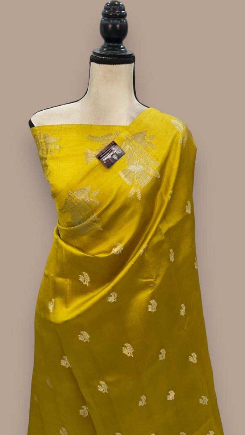 Puede incluir: Un sari de seda amarillo con un patr&oacute;n floral dorado. El sari est&aacute; drapeado en un maniqu&iacute;.
