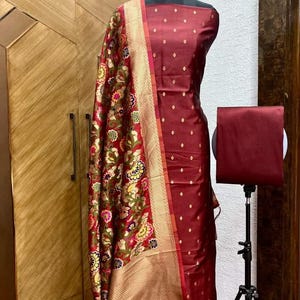 May include: A maroon dress fabric with a small diamond pattern, a maroon dupatta with a floral pattern, and a maroon plain fabric. The fabric is displayed on a mannequin.
