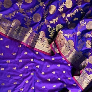 Könnte beinhalten: Ein lebendiger violetter Stoff mit goldenen Blumenmustern und Blattakzenten. Der Stoff hat aufwendige goldene Bordüren mit rotem Besatz. Das Design ist reichhaltig und detailliert, geeignet für Kleidung oder Heimdekoration.