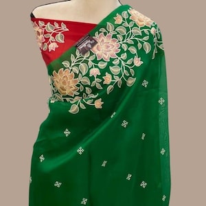 Op de afbeelding: Een groene sari met bloemenborduurwerk en een rode blouse. De sari heeft ingewikkelde bloempatronen in tinten goud en wit langs de rand. De blouse is effen rood van kleur, wat een opvallend contrast vormt met de groene stof.