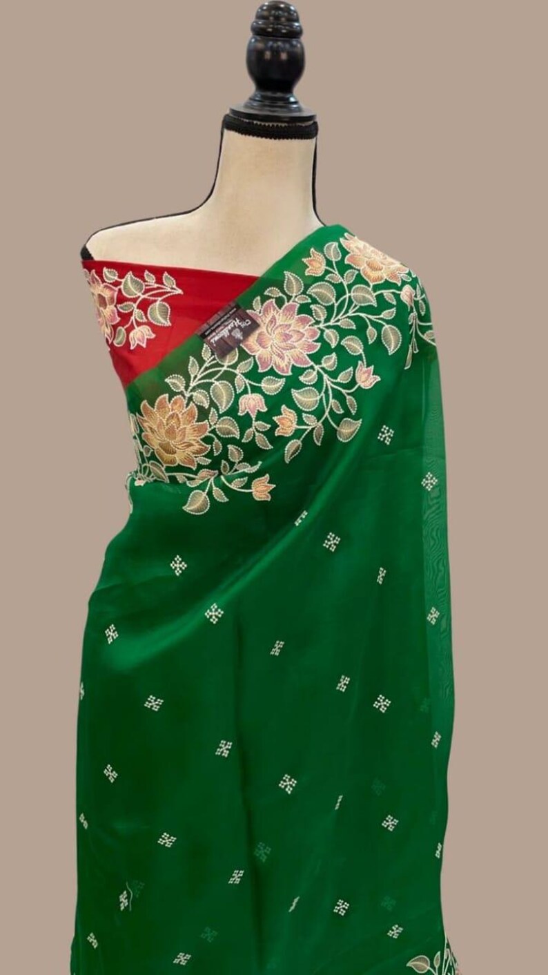 Op de afbeelding: Een groene sari met bloemenborduurwerk en een rode blouse. De sari heeft ingewikkelde bloempatronen in tinten goud en wit langs de rand. De blouse is effen rood van kleur, wat een opvallend contrast vormt met de groene stof.