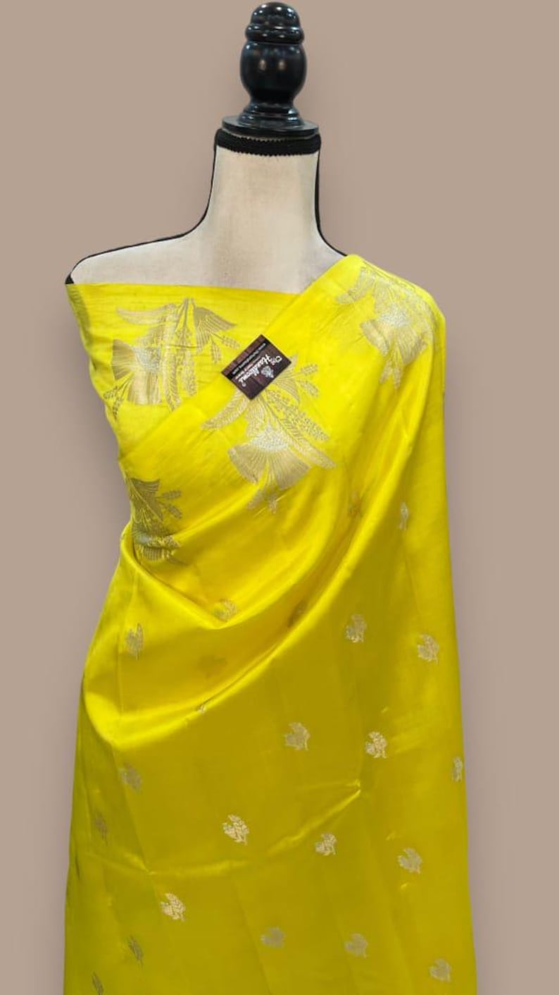 Puede incluir: Un sari de seda amarillo con un patr&oacute;n floral plateado. El sari est&aacute; drapeado sobre un maniqu&iacute;.