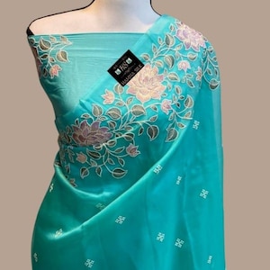 Op de afbeelding: Een turquoise sari met bloemenborduurwerk in roze, groen en goud. De sari heeft een delicaat patroon van kleine wit geborduurde vierkantjes. De stof is over een mannequin gedrapeerd.