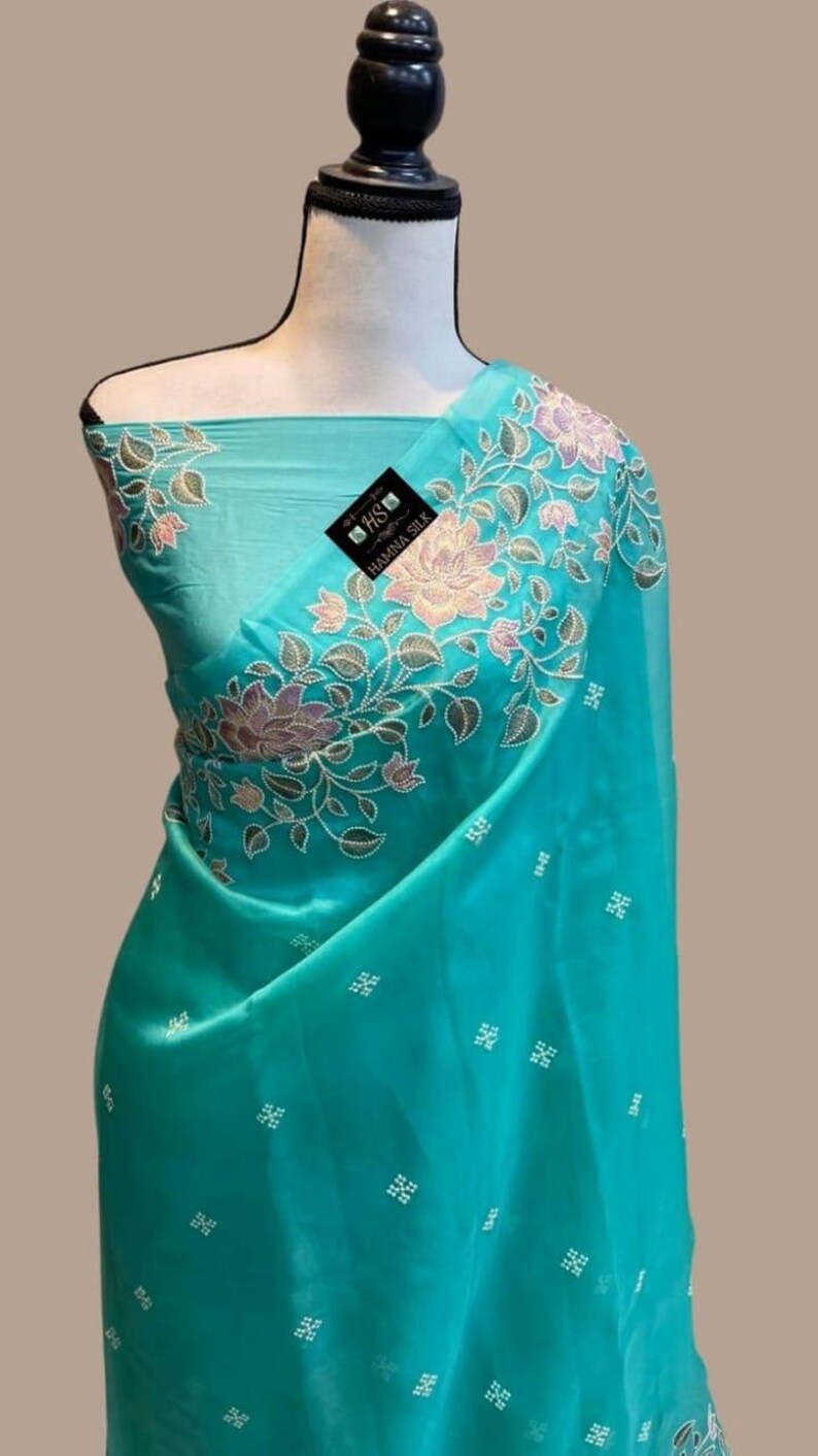 Op de afbeelding: Een turquoise sari met bloemenborduurwerk in roze, groen en goud. De sari heeft een delicaat patroon van kleine wit geborduurde vierkantjes. De stof is over een mannequin gedrapeerd.