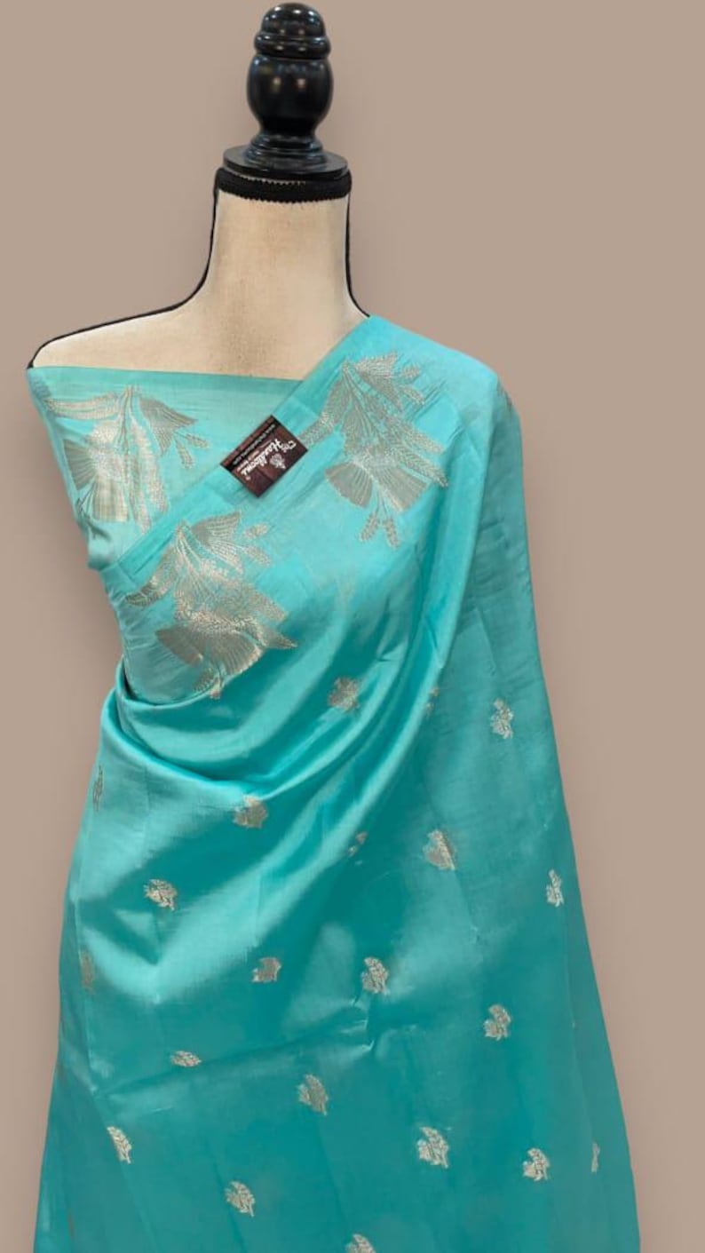 Puede incluir: Un sari de seda azul turquesa con bordados plateados. El sari tiene un dise&ntilde;o floral y est&aacute; drapeado en un maniqu&iacute;.