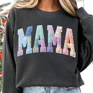 Op de afbeelding: Donkergrijze sweatshirt met het woord "MAMA" op de borst. De letters zijn gemaakt van patchwork stof in verschillende pastelkleuren en bloempatronen. Een comfortabele en stijlvolle top.
