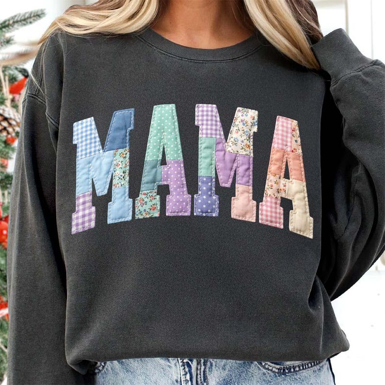Op de afbeelding: Donkergrijze sweatshirt met het woord "MAMA" op de borst. De letters zijn gemaakt van patchwork stof in verschillende pastelkleuren en bloempatronen. Een comfortabele en stijlvolle top.