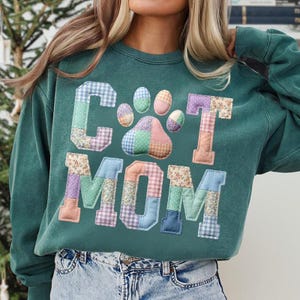 Pode incluir: Uma camisola verde-azulado com as palavras "CAT MOM" em letras de patchwork e um desenho de pegada. As letras e a pegada s&atilde;o feitas de v&aacute;rios tecidos estampados em cores pastel. A camisola tem mangas compridas e gola redonda.