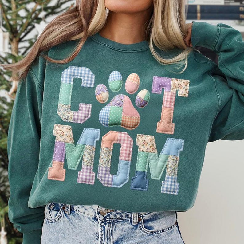 Pode incluir: Uma camisola verde-azulado com as palavras "CAT MOM" em letras de patchwork e um desenho de pegada. As letras e a pegada s&atilde;o feitas de v&aacute;rios tecidos estampados em cores pastel. A camisola tem mangas compridas e gola redonda.