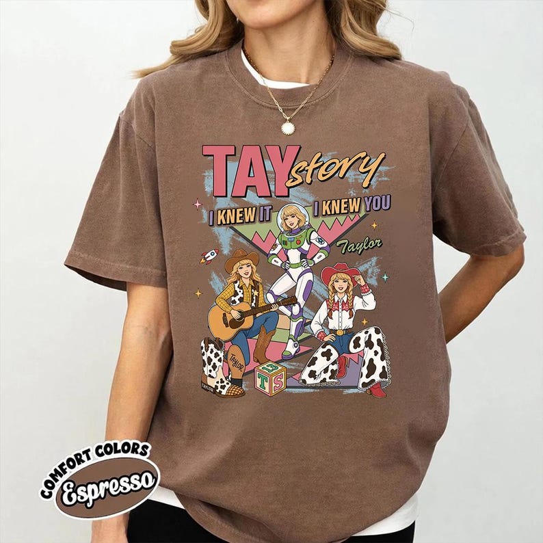 May include: A brown short-sleeve t-shirt featuring a graphic print with the text TAYstory I KNEW IT I KNEW YOU Taylor. The design includes three illustrated figures in cowboy and space-themed outfits, a guitar, a toy block, and stars.
