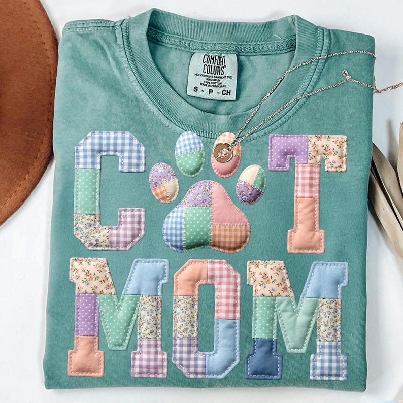 Pode incluir: Uma camiseta Comfort Colors turquesa com as palavras "CAT MOM" aplicadas em tecido patchwork. O design inclui uma pegada de pata e padr&otilde;es florais, xadrez e lisos. Um colar de prata com um pingente repousa na camisa.