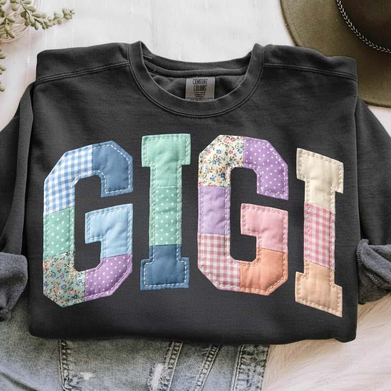 Op de afbeelding: Een donkergrijze sweatshirt met de naam "GIGI" in grote letters in patchwork-stijl. De letters zijn gemaakt van verschillende stoffen met patronen, waaronder ruitjes, bloemen en stippen. De sweatshirt heeft een ronde hals en lange mouwen.
