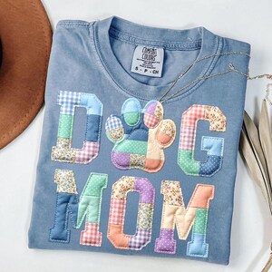 以下が含まれることがあります： 「Dog Mom」と足跡のパッチワークデザインが施された、青色のComfort Colors Tシャツ。文字と足跡は、青、緑、ピンク、オレンジの色合いの様々な柄の生地で作られています。金のネックレスがシャツの上にかけられています。