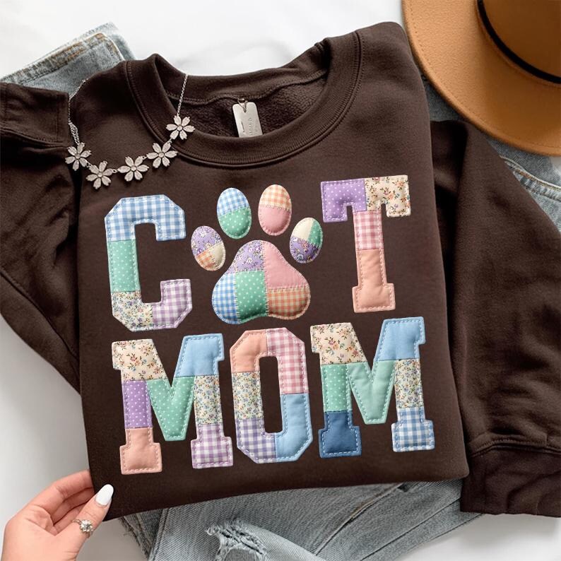 Pode incluir: Um moletom marrom com as palavras "Cat Mom" em letras de patchwork e uma pegada. As letras e a pegada s&atilde;o feitas de v&aacute;rios tecidos estampados em cores pastel. Um colar de prata com pingentes de flores est&aacute; drapeado no moletom.
