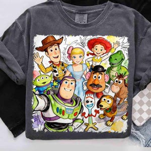 Pode incluir: Uma camiseta cinza carv&atilde;o com um gr&aacute;fico em estilo aquarela de personagens do filme Toy Story. O design inclui Woody, Buzz Lightyear, Bo Peep e outros personagens. O gr&aacute;fico &eacute; emoldurado com uma borda branca.
