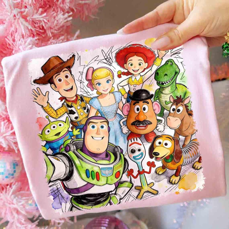 Pode incluir: Uma camiseta rosa claro com uma ilustra&ccedil;&atilde;o colorida de personagens de Toy Story. O design apresenta Woody, Buzz Lightyear e outros personagens do filme. A obra de arte &eacute; em estilo aquarela.