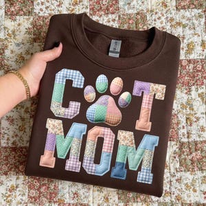 Pode incluir: Um moletom marrom com as palavras "CAT MOM" em letras de patchwork e um design de pegada. As letras e a pegada s&atilde;o feitas de v&aacute;rios tecidos estampados em cores pastel. O moletom est&aacute; dobrado e &eacute; segurado por uma pessoa.