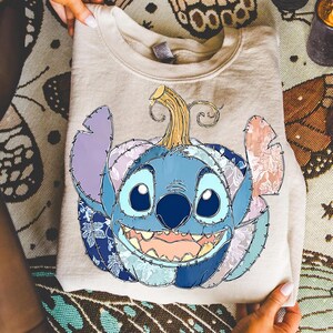 May include: A beige sweatshirt featuring a Stitch-themed pumpkin design. The pumpkin is blue with patchwork details in various shades of blue, purple, and pink. The design includes a smiling face and a curly stem.