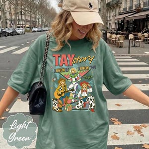 May include: A sage green short-sleeve t-shirt featuring a graphic print with the text TAYstory I KNEW IT I KNEW YOU. The design includes three illustrated figures in cowboy and space-themed outfits, a guitar, and a block with the letters TS.