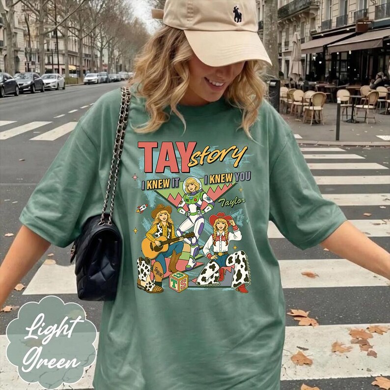 May include: A sage green short-sleeve t-shirt featuring a graphic print with the text TAYstory I KNEW IT I KNEW YOU. The design includes three illustrated figures in cowboy and space-themed outfits, a guitar, and a block with the letters TS.