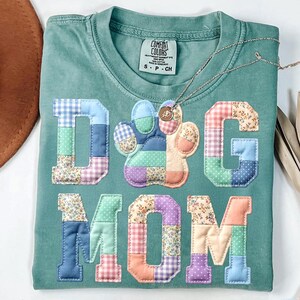 以下が含まれることがあります： 鮮やかなパッチワーク生地で「DOG MOM」の文字がアップリケされた、ティール色のComfort Colors Tシャツ。 「O」は足跡です。 小さなペンダントが付いたシルバーのネックレスがシャツの上にあります。