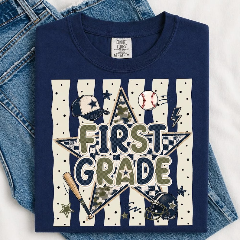 以下が含まれることがあります： FIRST GRADEという文字がプリントされたネイビーブルーのTシャツ。大きな星、野球帽、野球ボール、バット、フットボールヘルメットが描かれており、背景には黒いドットが入ったクリーム色の縦縞模様があしらわれています。