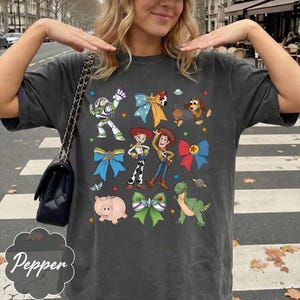Op de afbeelding: Een antracietgrijs T-shirt met korte mouwen en een grafische print van Toy Story-personages, waaronder Buzz Lightyear, Woody, Jessie, Slinky Dog, Rex en Hamm, afgewisseld met kleurrijke strikken en kleine ster- en planeeticonen.