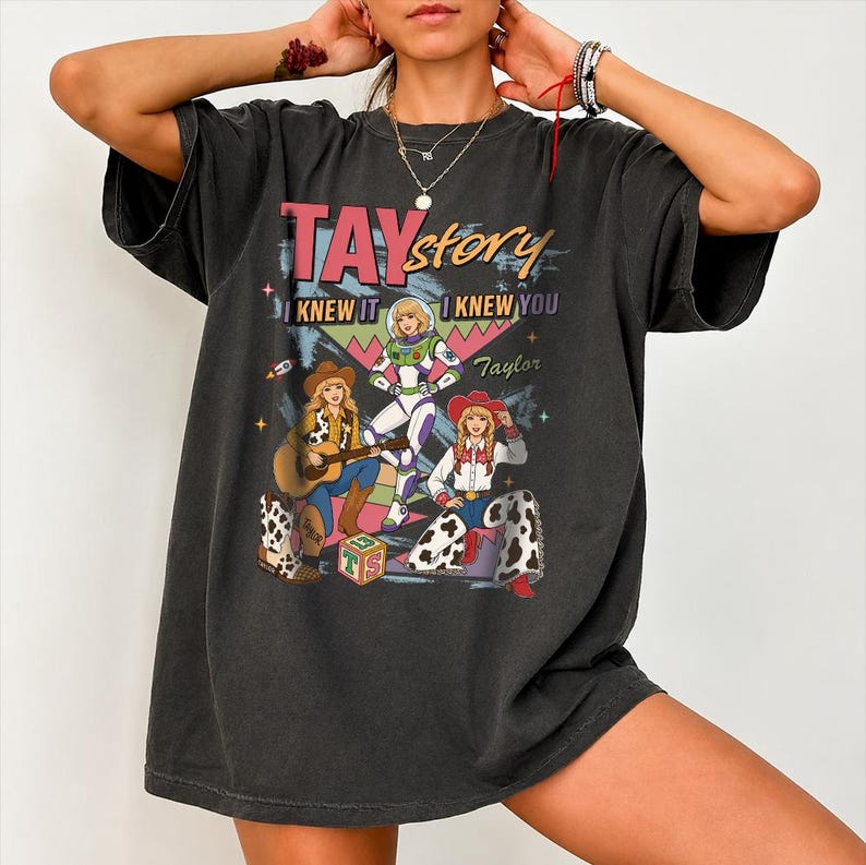 May include: A charcoal gray short-sleeve t-shirt featuring a graphic print with the text TAYstory, I KNEW IT, I KNEW YOU, and Taylor. The design includes three illustrated figures in cowboy and space-themed outfits, a guitar, and a toy block.