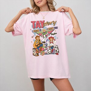 May include: A light pink t-shirt featuring a graphic print with the text TAYstory, I KNEW IT, I KNEW YOU, and Taylor. The design includes illustrations of three figures in cowboy-inspired outfits, a space-themed character, and a toy block.