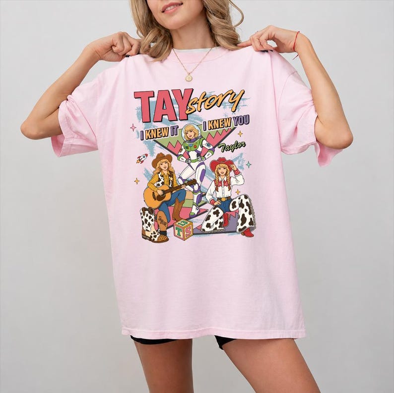 May include: A light pink t-shirt featuring a graphic print with the text TAYstory, I KNEW IT, I KNEW YOU, and Taylor. The design includes illustrations of three figures in cowboy-inspired outfits, a space-themed character, and a toy block.