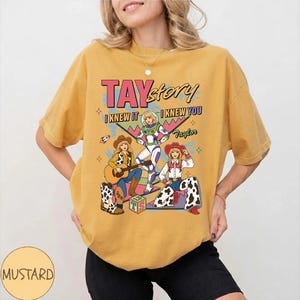 May include: A mustard yellow short-sleeve t-shirt featuring a graphic print with the text TAYstory, I KNEW IT, I KNEW YOU, and Taylor. The design depicts three illustrated figures in cowboy and space-themed outfits with stars and blocks.