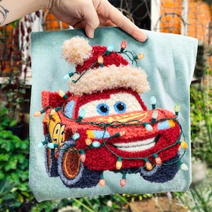 Peut inclure: Un motif de voiture rouge Lightning McQueen avec un bonnet de Père Noël et des guirlandes lumineuses sur un fond bleu clair. La voiture a des yeux bleus et une bouche souriante. Le numéro 96 est visible sur le côté de la voiture. Le motif est sur un coussin carré.