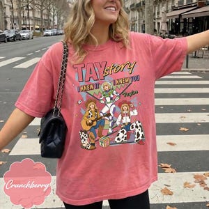 May include: A coral-colored short-sleeve t-shirt featuring a graphic print with the text TAYstory, I KNEW IT, I KNEW YOU, and Taylor. The design includes three illustrated figures in cowboy and space-themed outfits, a guitar, and a block with TS.