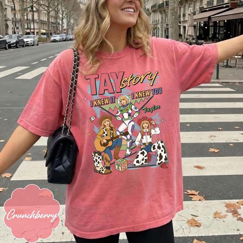 May include: A coral-colored short-sleeve t-shirt featuring a graphic print with the text TAYstory, I KNEW IT, I KNEW YOU, and Taylor. The design includes three illustrated figures in cowboy and space-themed outfits, a guitar, and a block with TS.