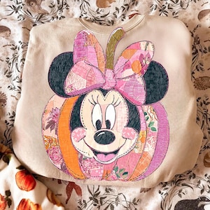 Puede incluir: Sudadera beige con un diseño de calabaza de Minnie Mouse. La calabaza está hecha de tela de patchwork en tonos rosas, naranjas y estampados florales. Las orejas de Minnie son negras y su lazo es rosa. El diseño está centrado en la parte delantera.