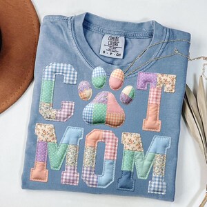 Pode incluir: Uma camiseta azul claro Comfort Colors com as palavras "CAT MOM" aplicadas em tecido patchwork. O design inclui uma pegada de pata de gato e letras em v&aacute;rios padr&otilde;es florais e xadrez. Um colar de ouro est&aacute; drapeado sobre a camisa.