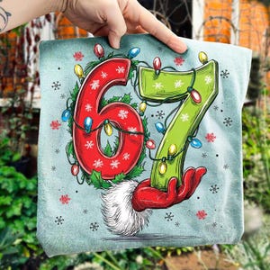 May include: A light blue Christmas-themed graphic featuring the numbers 67 in red and green, adorned with string lights and snowflakes. The number 6 is encircled by holly, and the number 7 is held by a red gloved hand.
