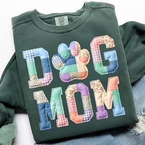以下が含まれることがあります： カラフルなパッチワーク文字と足跡で「DOG MOM」と書かれた濃い緑色のスウェットシャツ。文字と足跡は、花柄やギンガムチェックなど、さまざまな模様の生地で作られています。