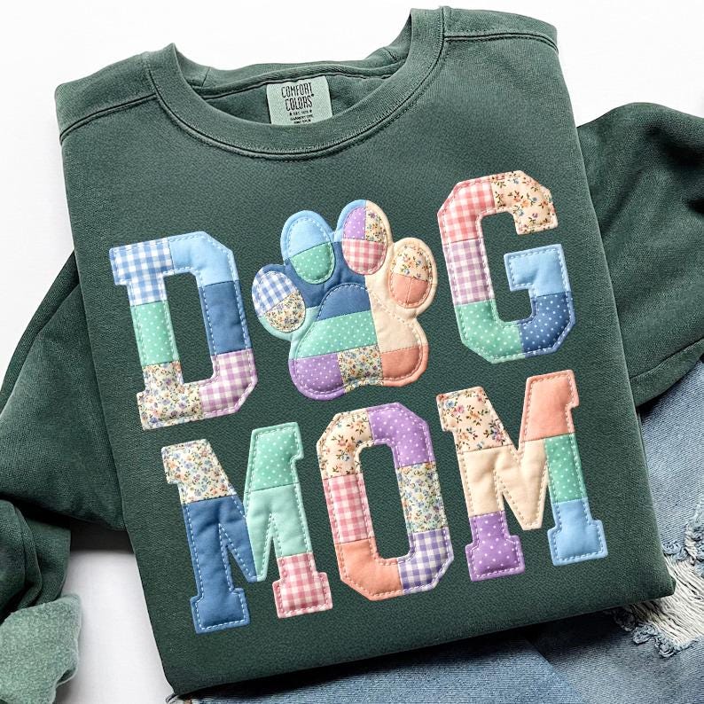 以下が含まれることがあります： カラフルなパッチワーク文字と足跡で「DOG MOM」と書かれた濃い緑色のスウェットシャツ。文字と足跡は、花柄やギンガムチェックなど、さまざまな模様の生地で作られています。